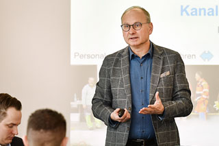 man with glasses, blue shirt and checked jacket lecturing